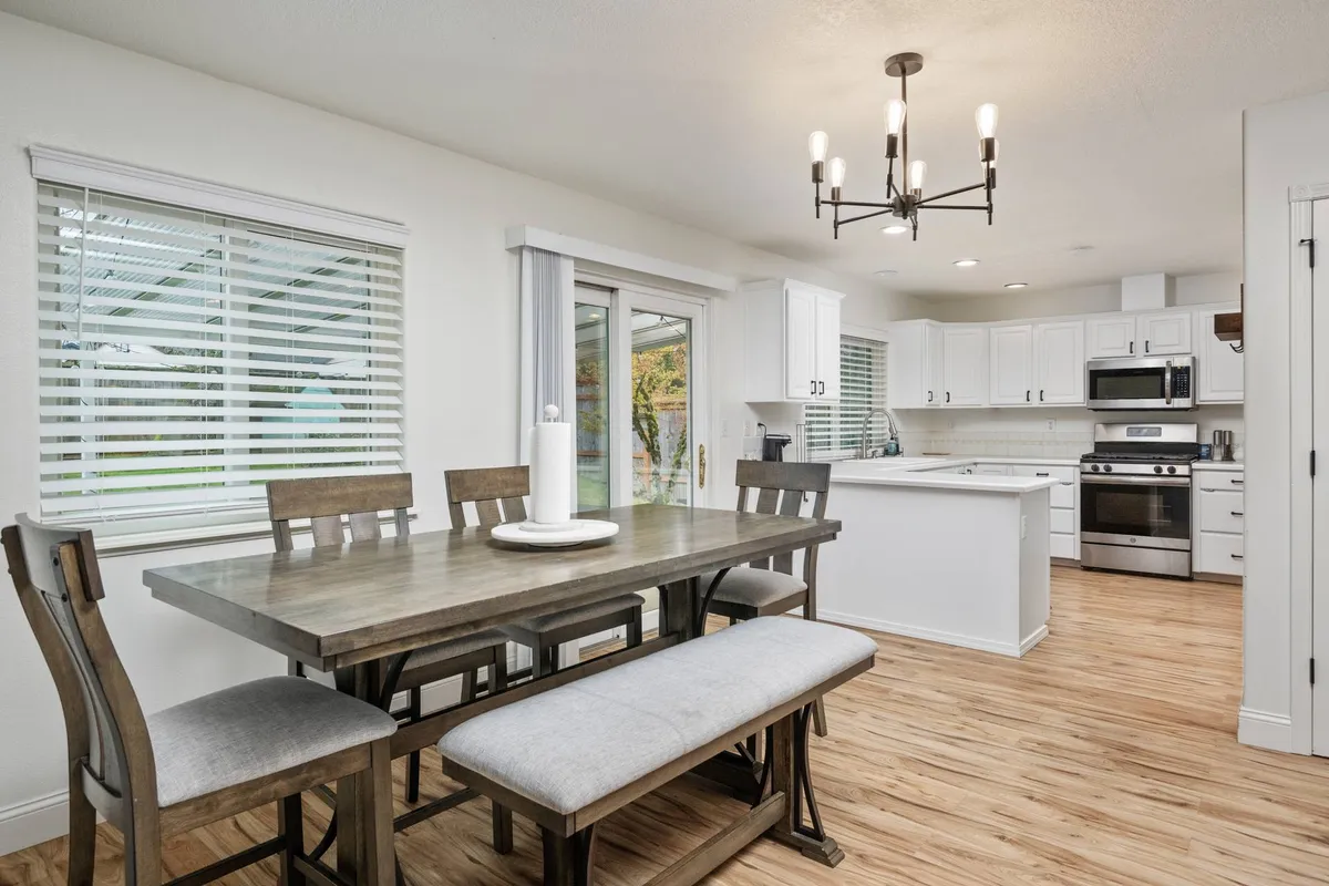 This image features a well-lit dining table with a stylish light fixture overhead and a kitchen island visible in the background. The arrangement suggests a seamless transition between the dining and kitchen areas, hinting at an open-concept layout ideal for gatherings and shared meals.