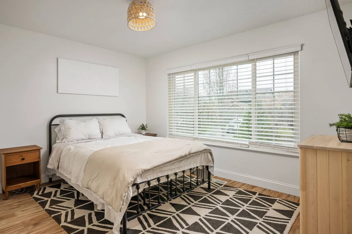 A serene bedroom with large windows allowing natural light to fill the space, a cozy rug, and a simple nightstand. The atmosphere is quiet, airy, and inviting, ideal for starting the day in tranquility.