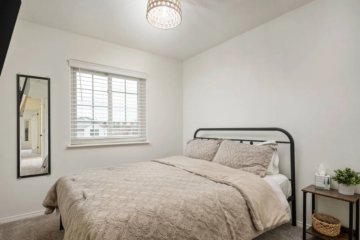A calm and inviting bedroom with a comfortable bed, a window streaming in morning light, a nightstand, and a mirror. The setting is cozy, peaceful, and ideal for restful sleep or quiet reading.